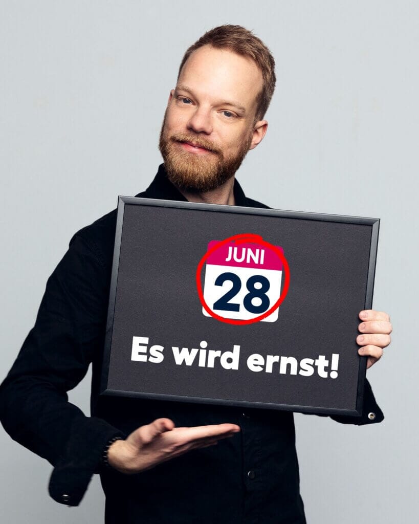 Ein Mann mit Bart und schwarzem Hemd hält ein Schild in die Kamera. Auf dem Schild steht groß: ‚28. Juni‘ mit Kalendergrafik, darüber der Schriftzug ‚Juni‘, rot eingekreist. Darunter: ‚Es wird ernst!‘. Er lächelt leicht und zeigt mit der Hand auf das Schild.