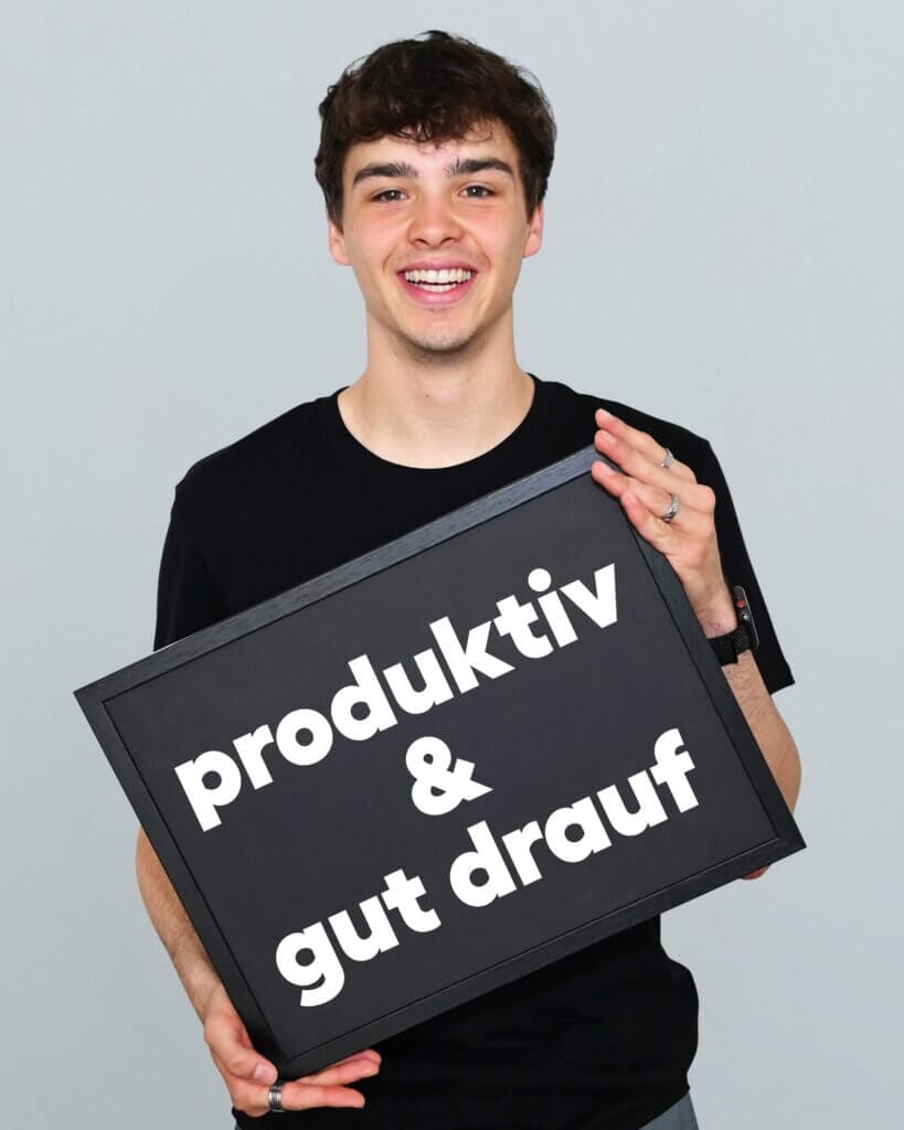 Ein junger Mann lächelt fröhlich und hält ein Schild mit der Aufschrift „produktiv & gut drauf“.