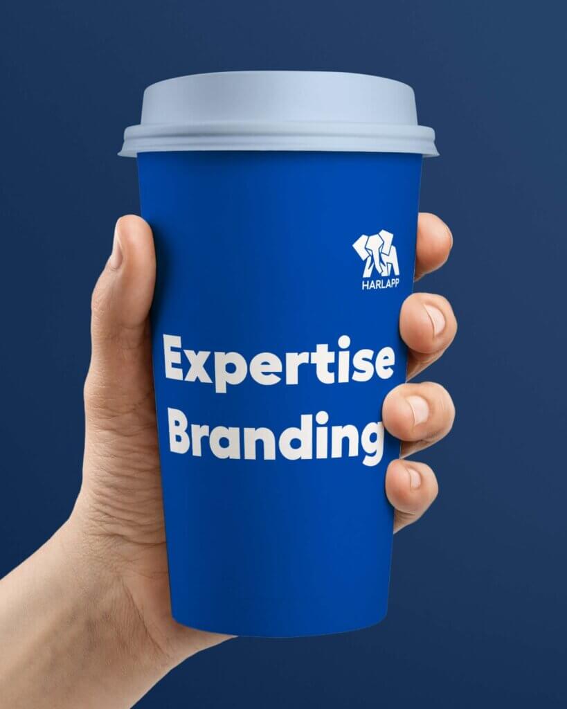 Eine Hand hält einen blauen Kaffeebecher mit dem Text "Expertise Branding" und dem HARLAPP-Logo.